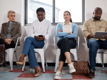 A waiting room with 4 people, they are waiting for a physiotherapy appointment. We can only see a small part of the room implying that it is very busy. One patient is on a tablet and one on a phone. Only one is smiling.