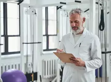 Physiotherapist takes notes in a gym on a clipboard, he is wearing a white tunic. in an article about evidence based practice in physiotherapy