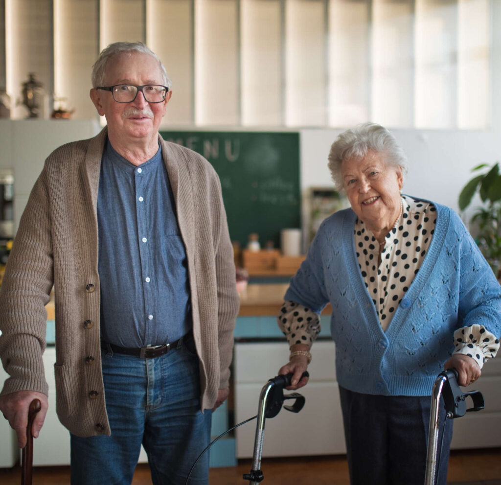 A senior man and woman walking with walker indoors in retirement nursing home. The image is in an article about whether physiotherapy could be conducted online for various patients.