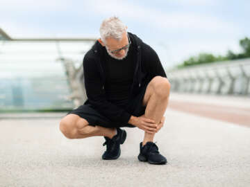 Senior man wearing black sportswear jogging outdoor by bridge, squating and touching his leg, appears to be suffering from shin splints. Image is in a blog about online physiotherapy treatment and shin splints.