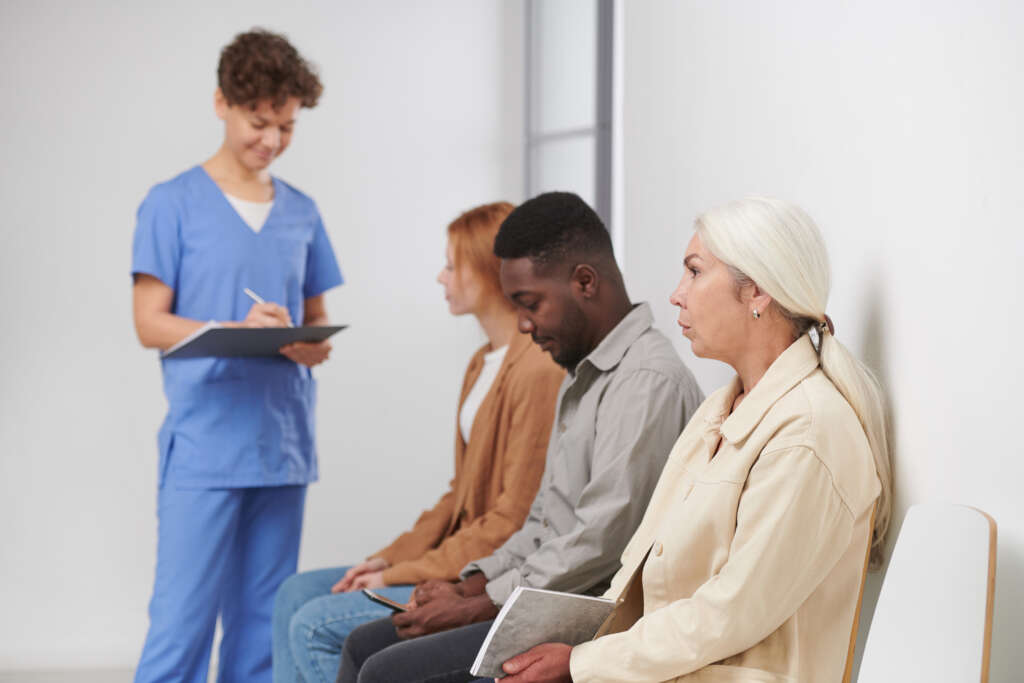 Group of three people sitting in queue waiting for consultation in modern hospital, medical worker standing near them making notes